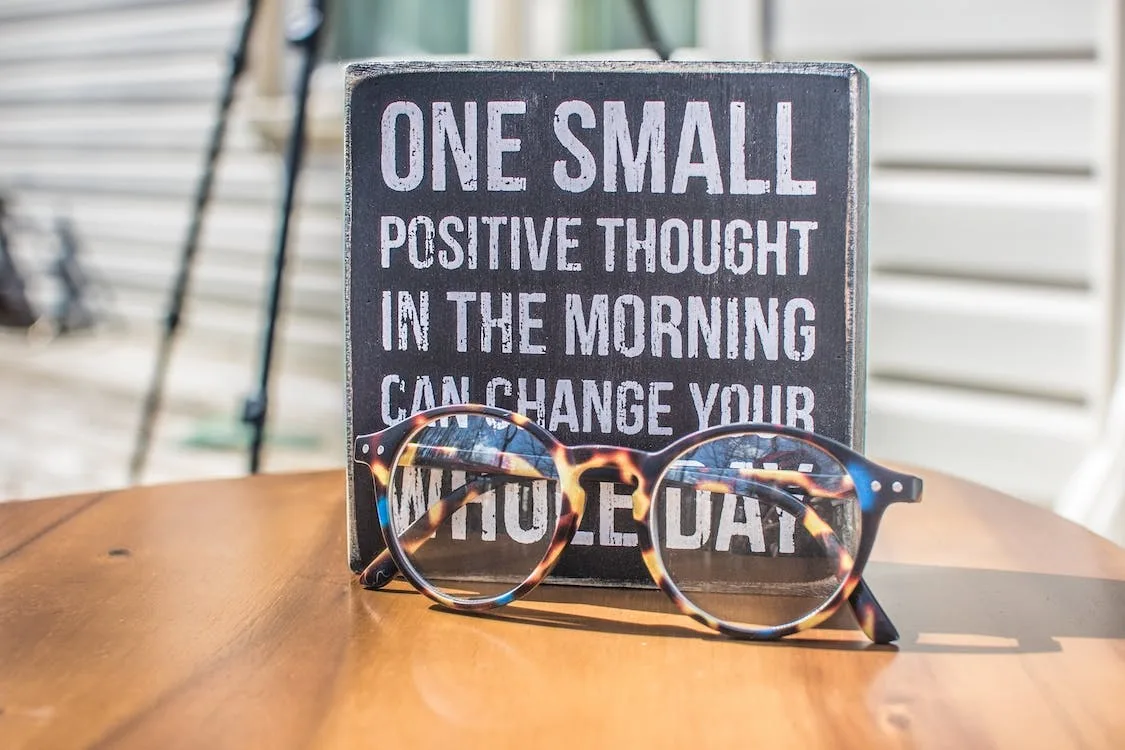 A wooden sign reading one small positive thought in the morning can change your whole day, with a pair of glasses in front