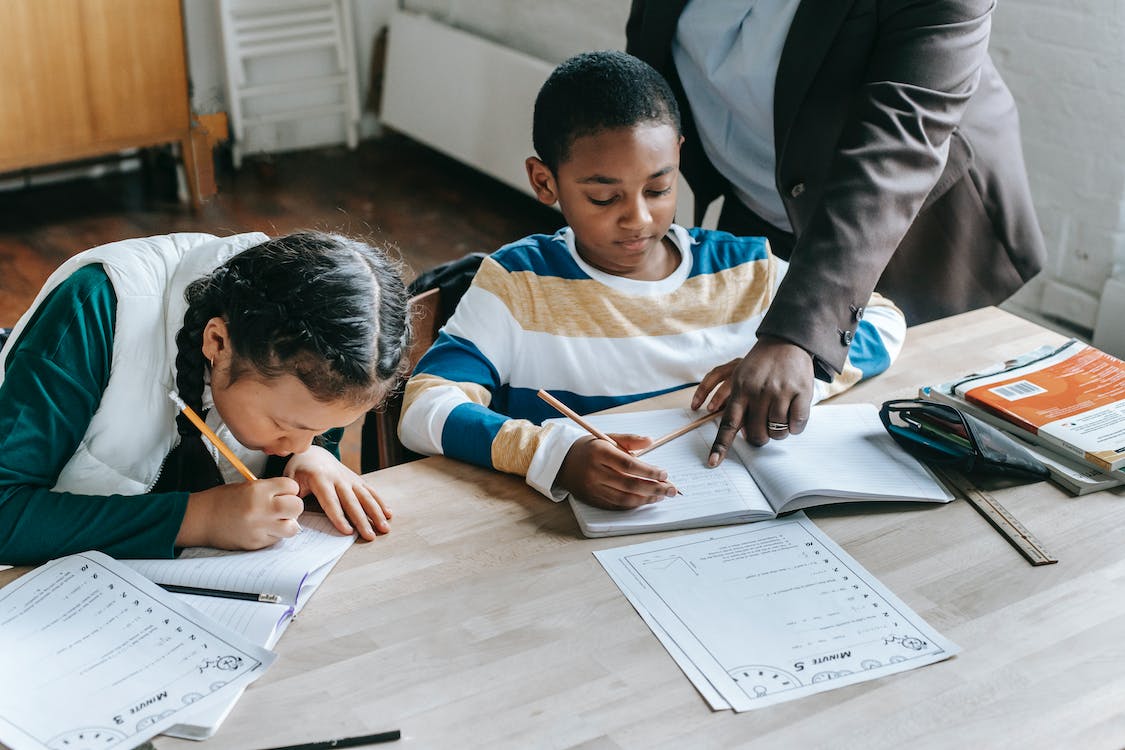 Free High angle of crop unrecognizable black female teacher explaining task to focused little schoolboy sitting at desk near attentive Asian classmate writing in notebook Stock Photo