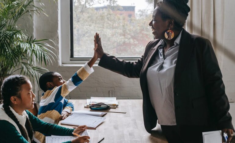 Free Side view of smiling black boy giving high five to teacher while sitting with diverse classmate girl at desk in classroom Stock Photo