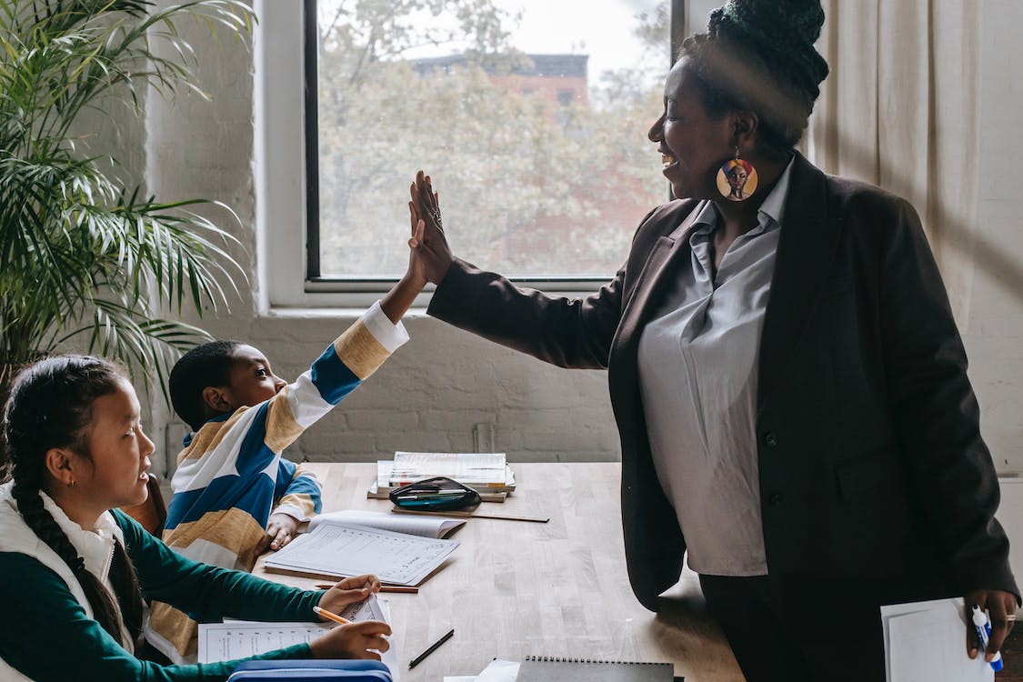 Free Side view of smiling black boy giving high five to teacher while sitting with diverse classmate girl at desk in classroom Stock Photo