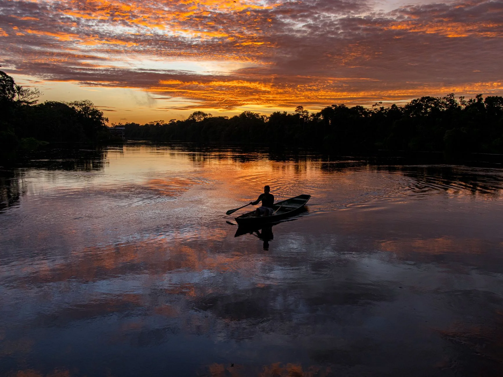 A fisherman rowing a small boat on the Amazon River at sunset with orange clouds reflected in the water