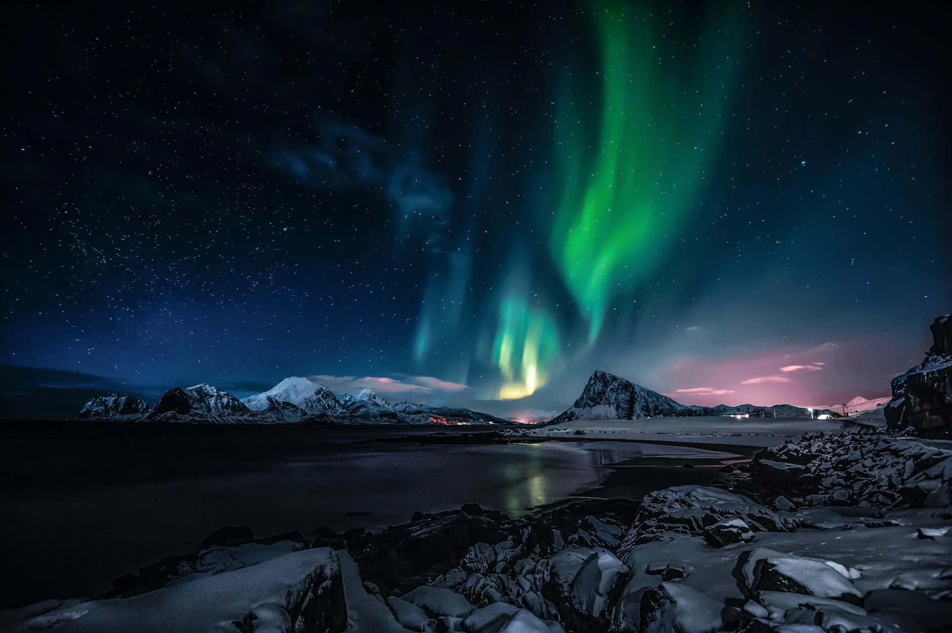 Green aurora curtains over snowy mountains and a dark shoreline under a starry sky in Nordland, Norway