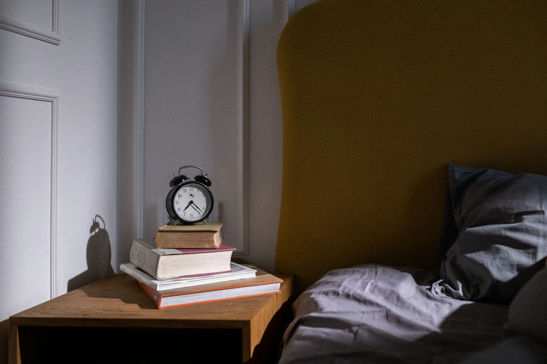 An alarm clock on a stack of books on a bedside table in a dim bedroom