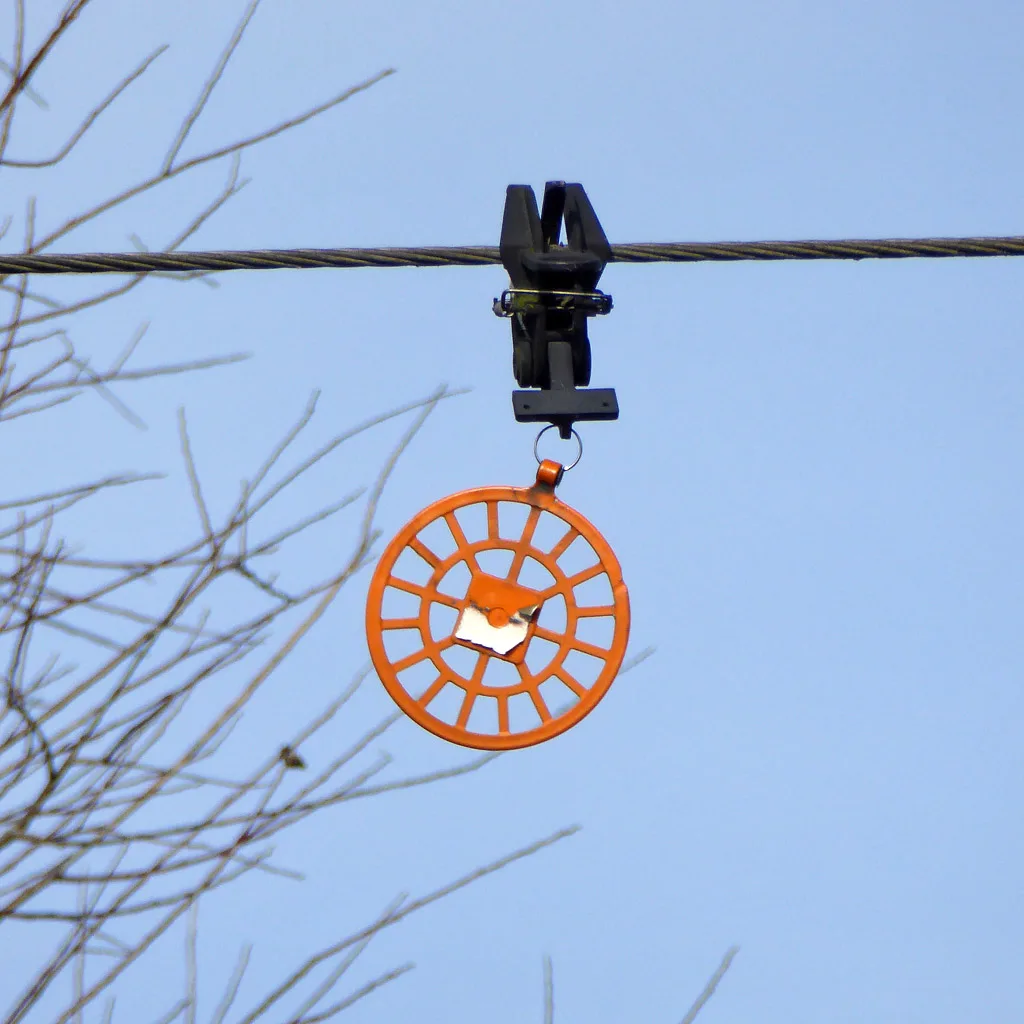 An orange spoked disc-shaped bird flight diverter hanging from a clamp on an overhead power line against a blue sky