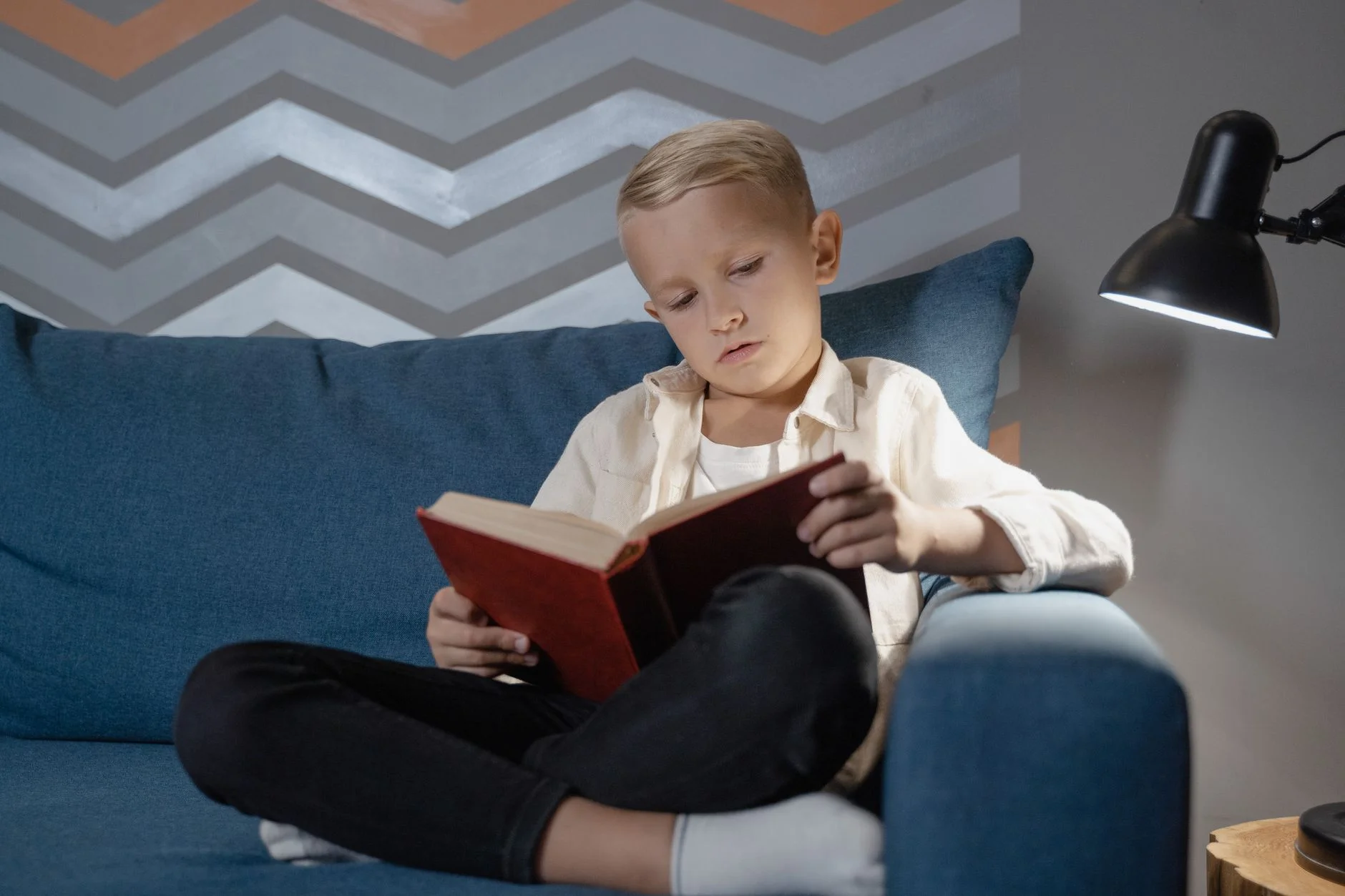 A boy sitting cross-legged on a blue sofa reading a book under a lamp