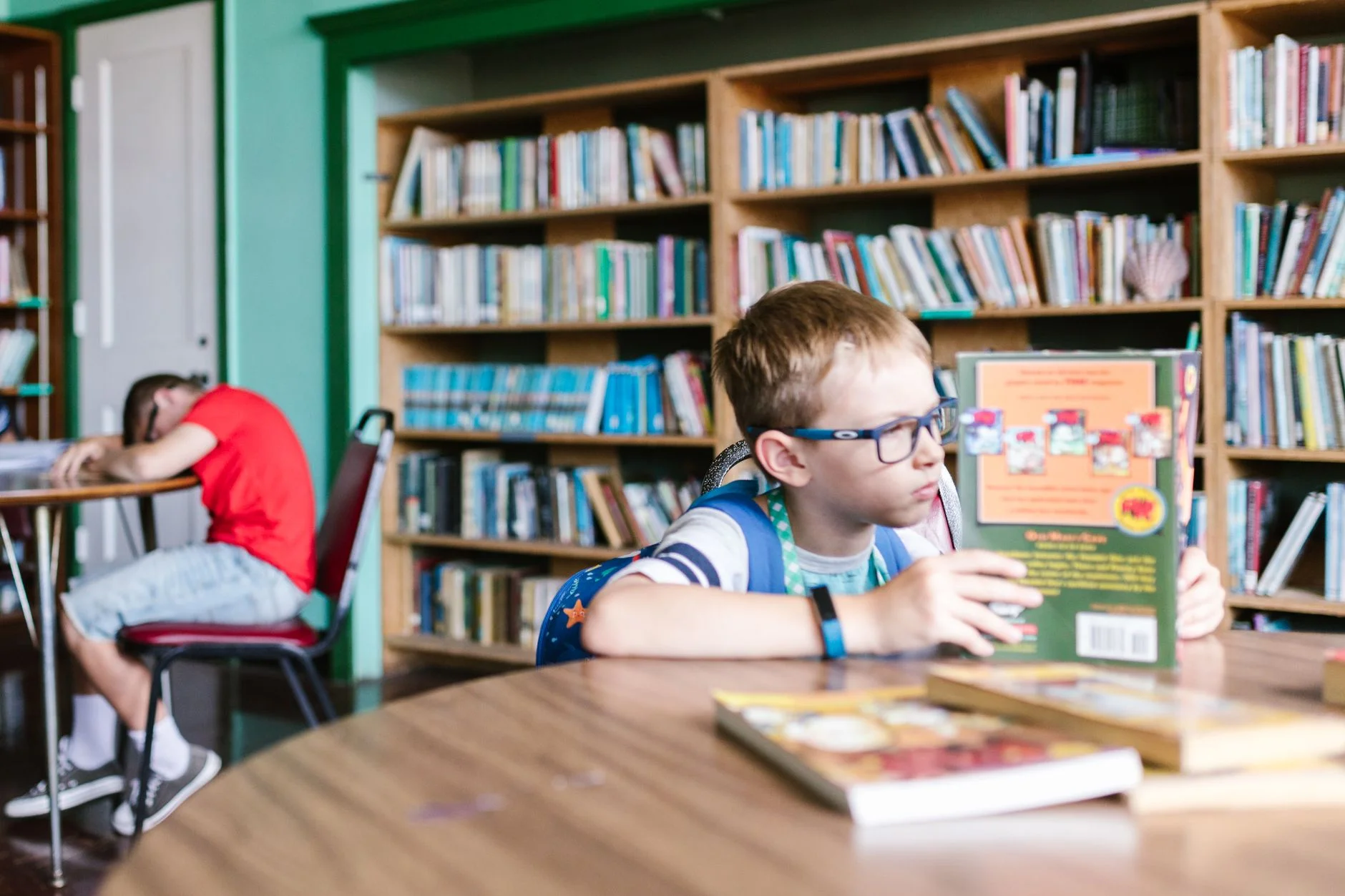 A boy with glasses reading at a table in a school library with shelves of books behind him