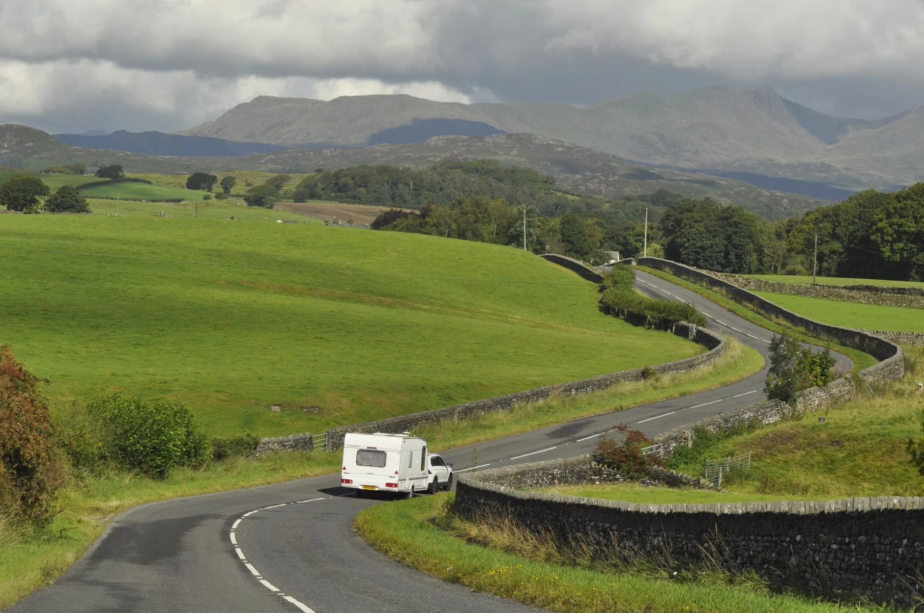 A car towing a white touring caravan along a narrow winding country road between stone walls and green hills