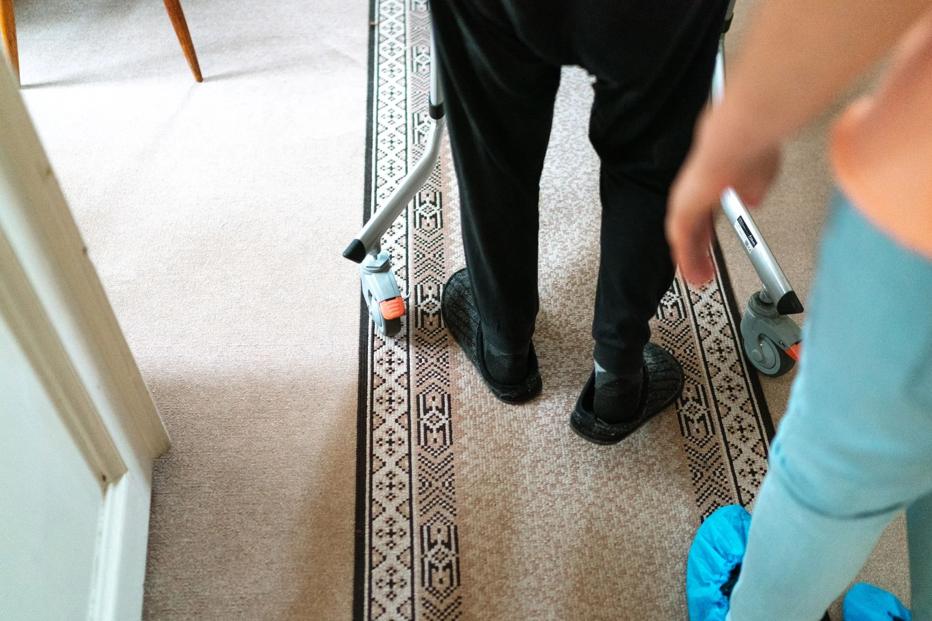 The legs of an older person in slippers walking with a walking frame along a carpeted hallway, while a carer in blue shoe covers walks close beside them