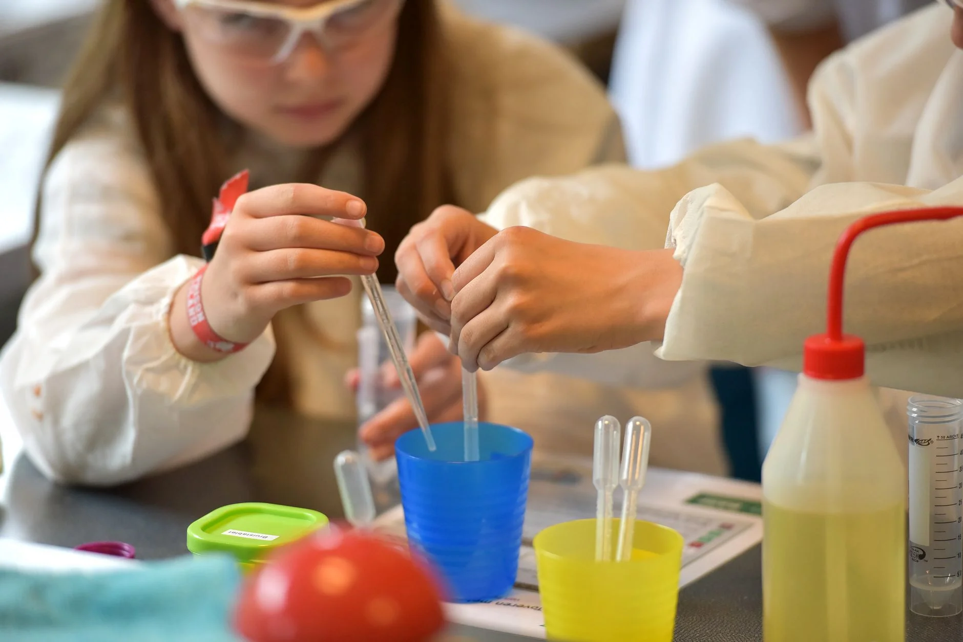 Two children wearing lab coats using plastic pipettes to move liquid into coloured cups