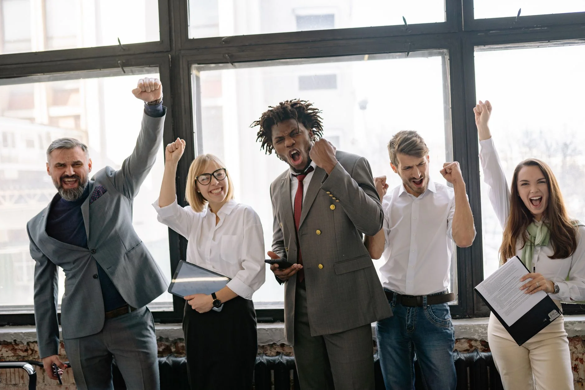 A group of five colleagues cheering with raised fists in front of large office windows