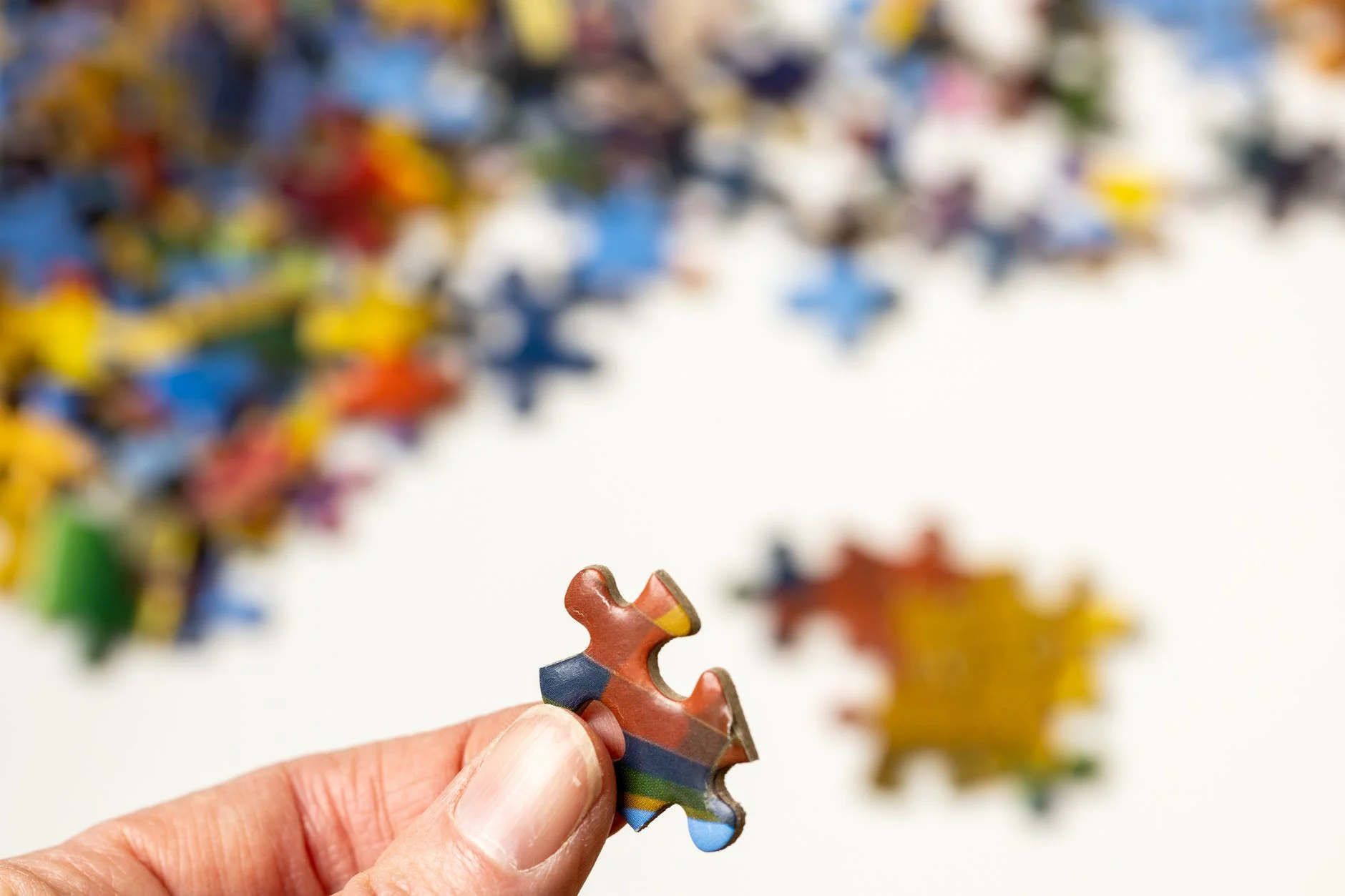 A hand holding up one colourful jigsaw puzzle piece in front of a blurred pile of other pieces