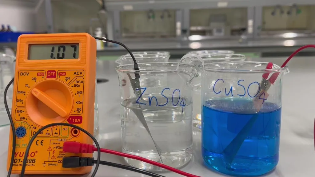 Daniell cell demonstration: zinc in a beaker of zinc sulfate and copper in a beaker of blue copper sulfate, joined by a salt bridge, with a multimeter reading 1.07