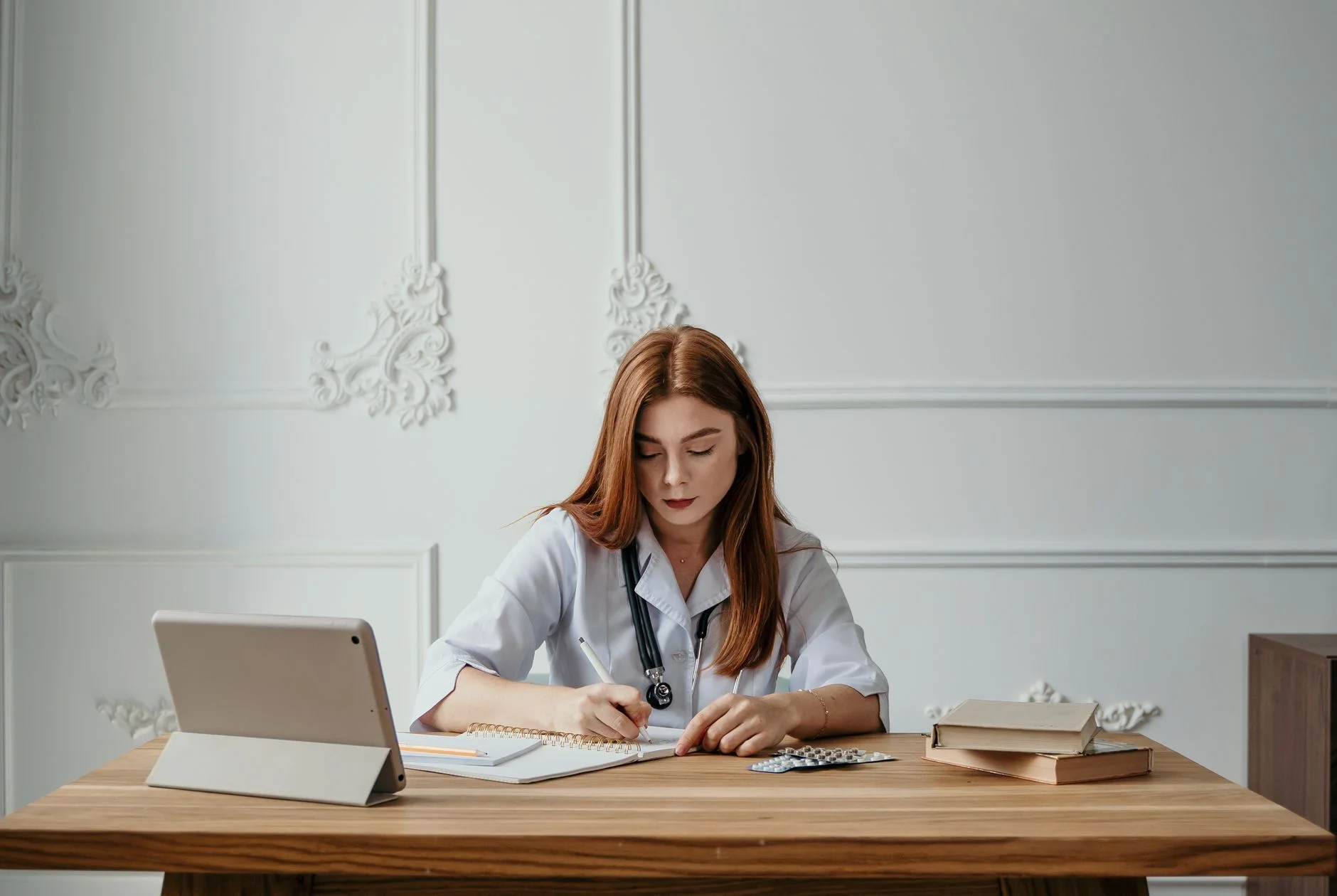 A doctor in a white coat writing notes at a desk with a tablet and books