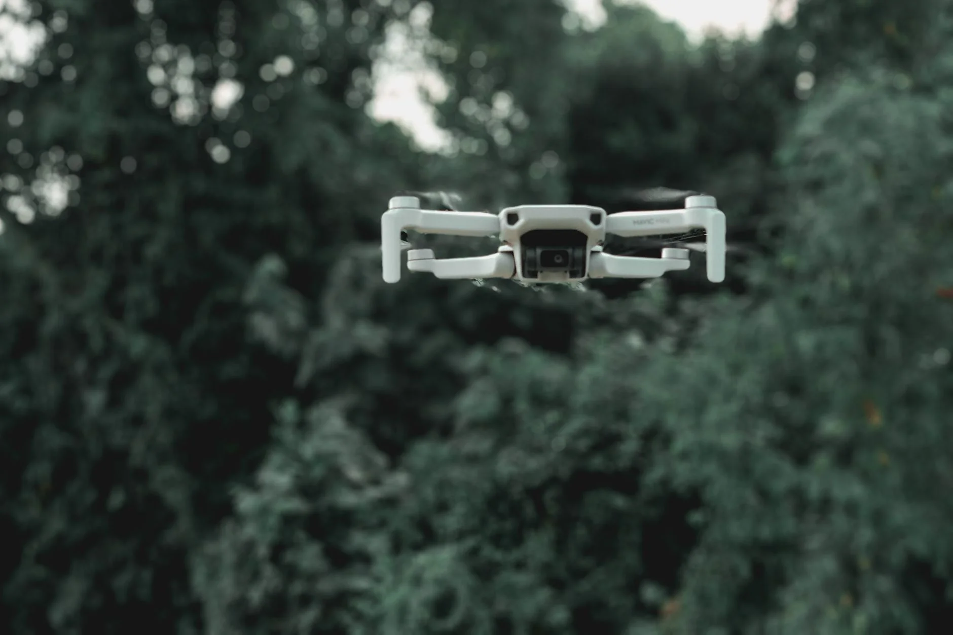 A small quadcopter drone with a camera hovering in front of green forest trees