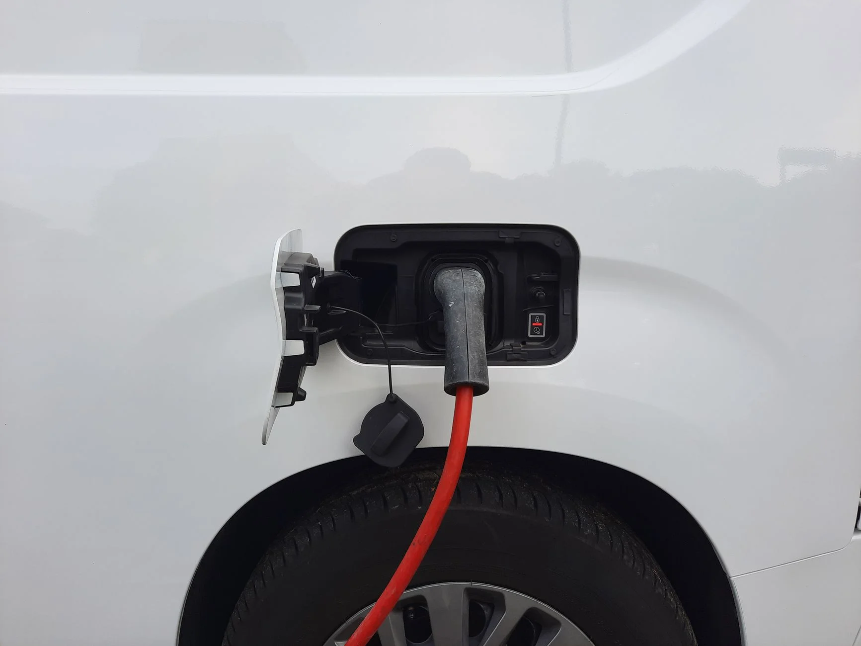 Red charging cable plugged into the open charge port on the side of a white car, above the wheel arch