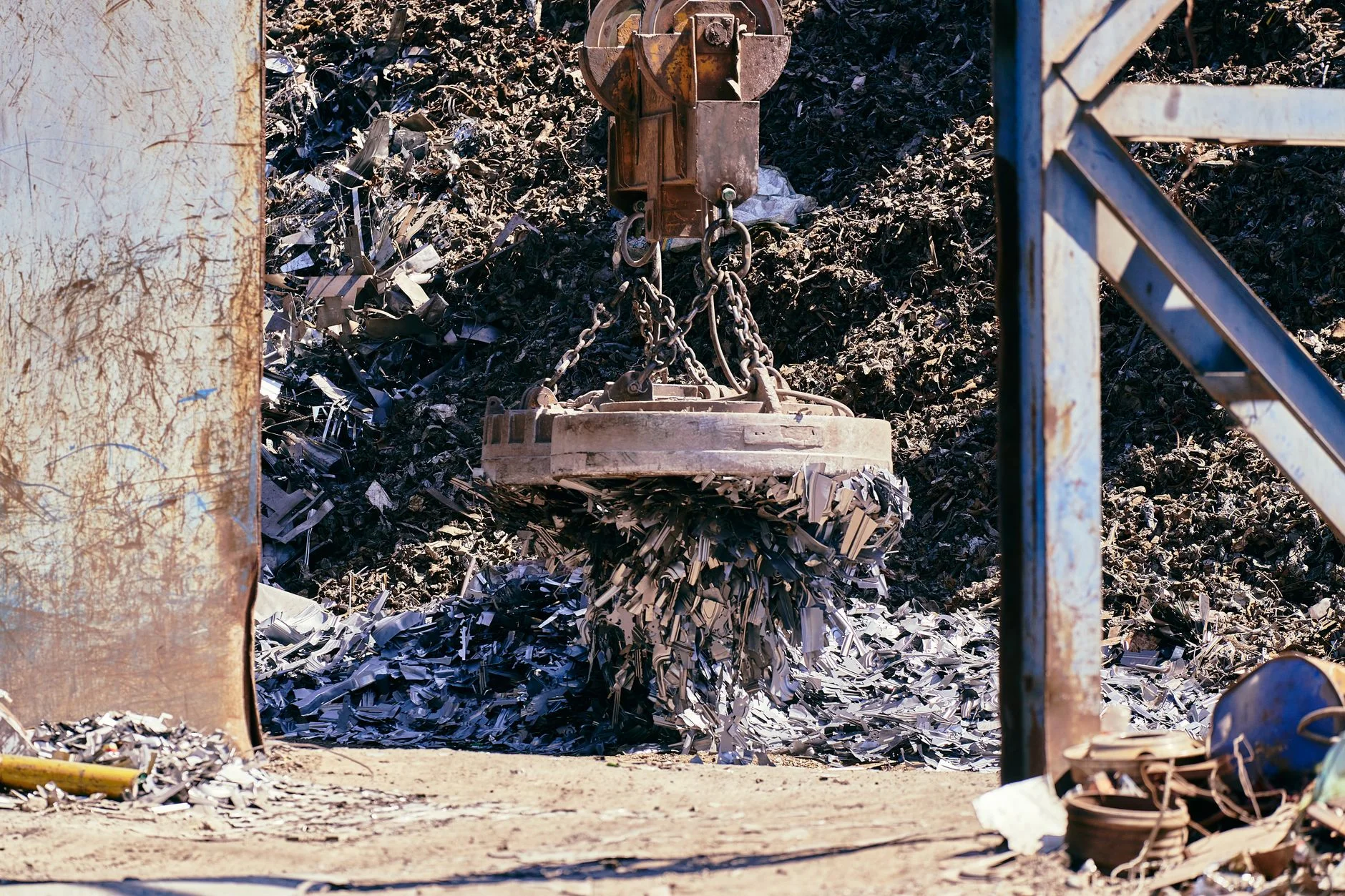 A large round electromagnet hanging from a crane chain, lifting a mass of shredded scrap steel in a recycling yard
