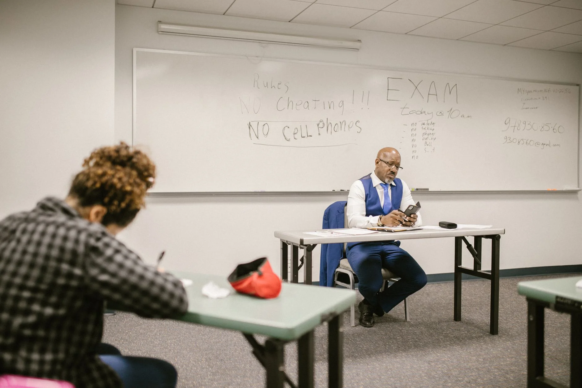 A proctor sits at a desk supervising a student taking an exam in a classroom
