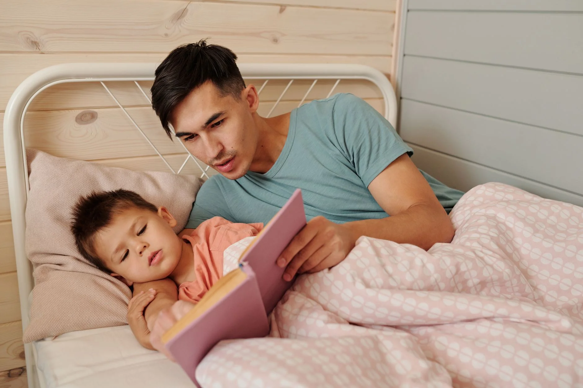 A father lying on a bed reading a picture book to his young son under a pink blanket