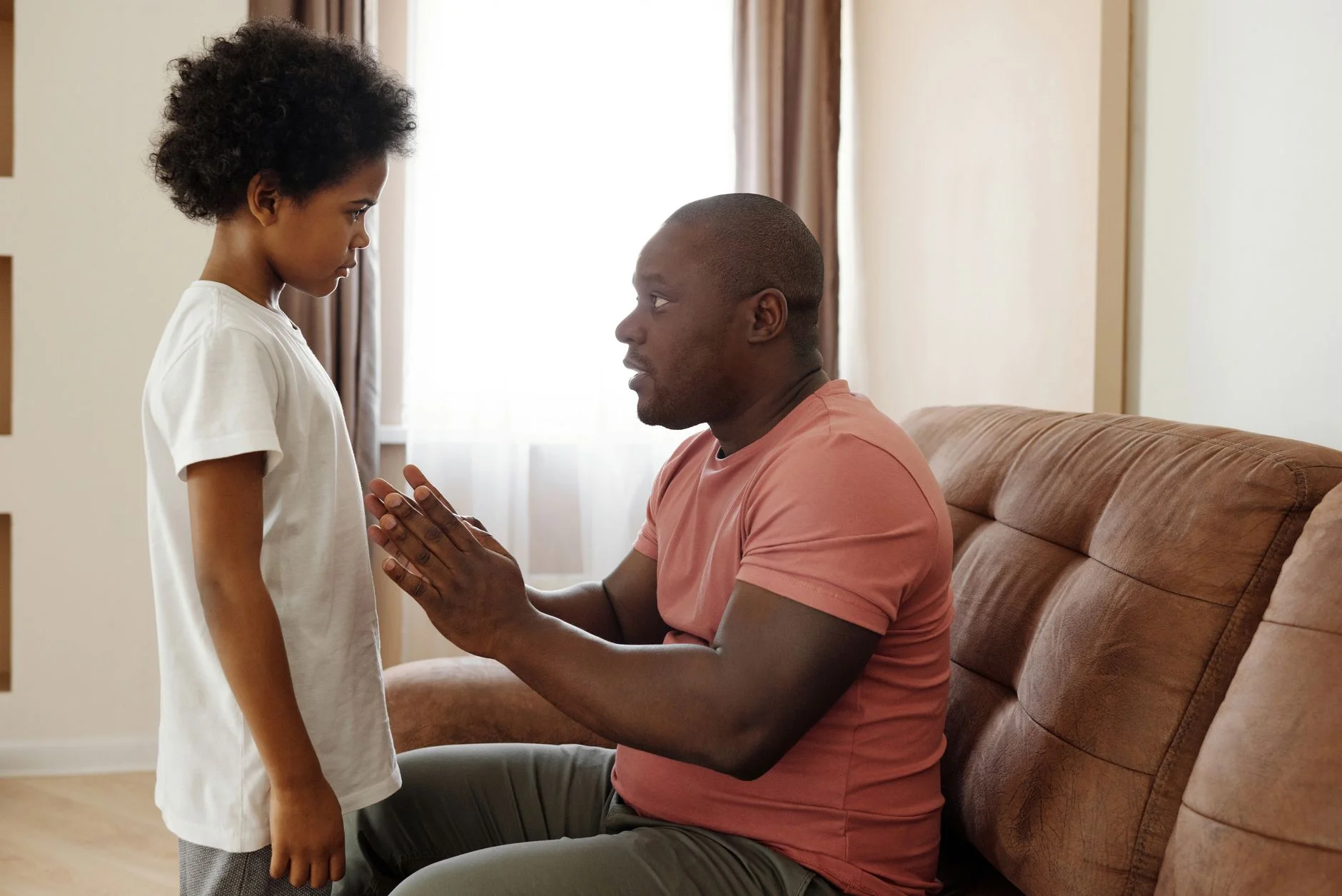 A father sitting on a sofa leaning towards his young son, who is standing in front of him, with his hands pressed together as he talks