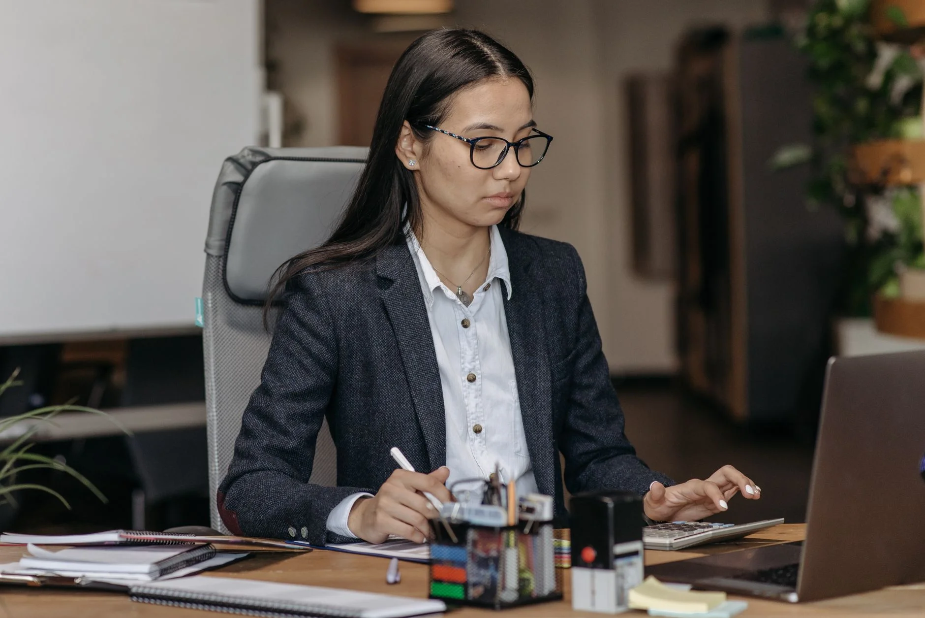 A woman in a blazer and glasses concentrating on her laptop at a tidy desk