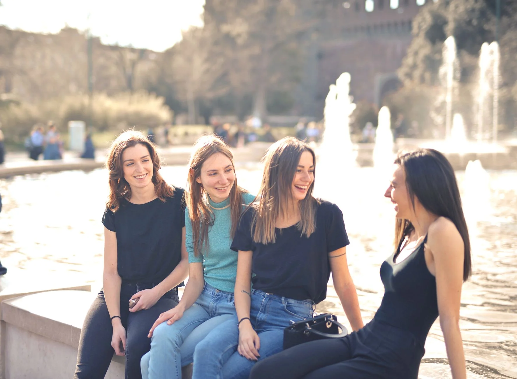 Four young women laughing together by a fountain on a sunny day