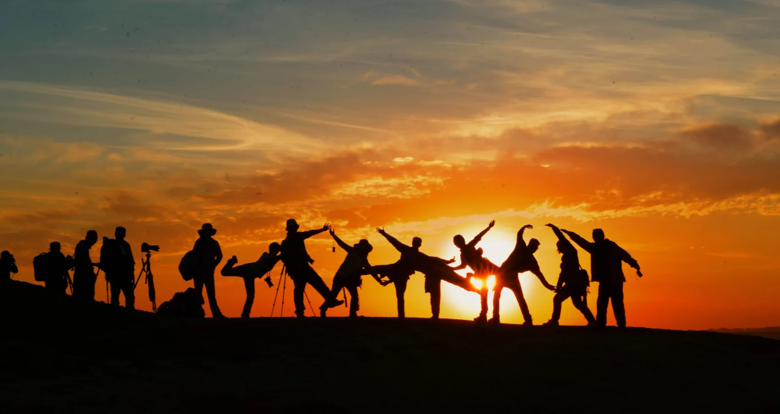 Silhouettes of a group of friends posing together on a hill against an orange sunset