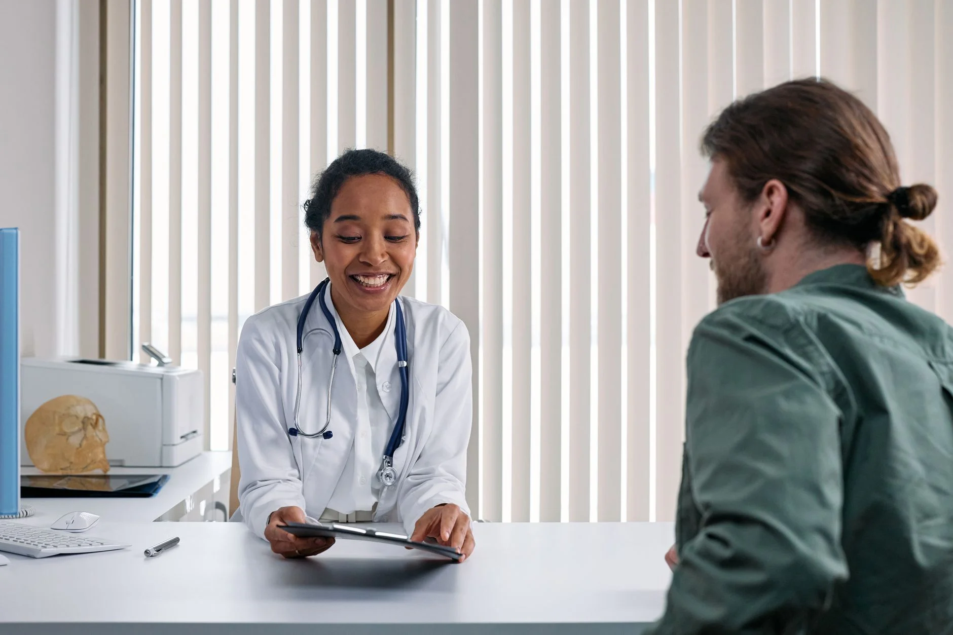 A smiling doctor talking with a patient across a desk in a bright clinic room