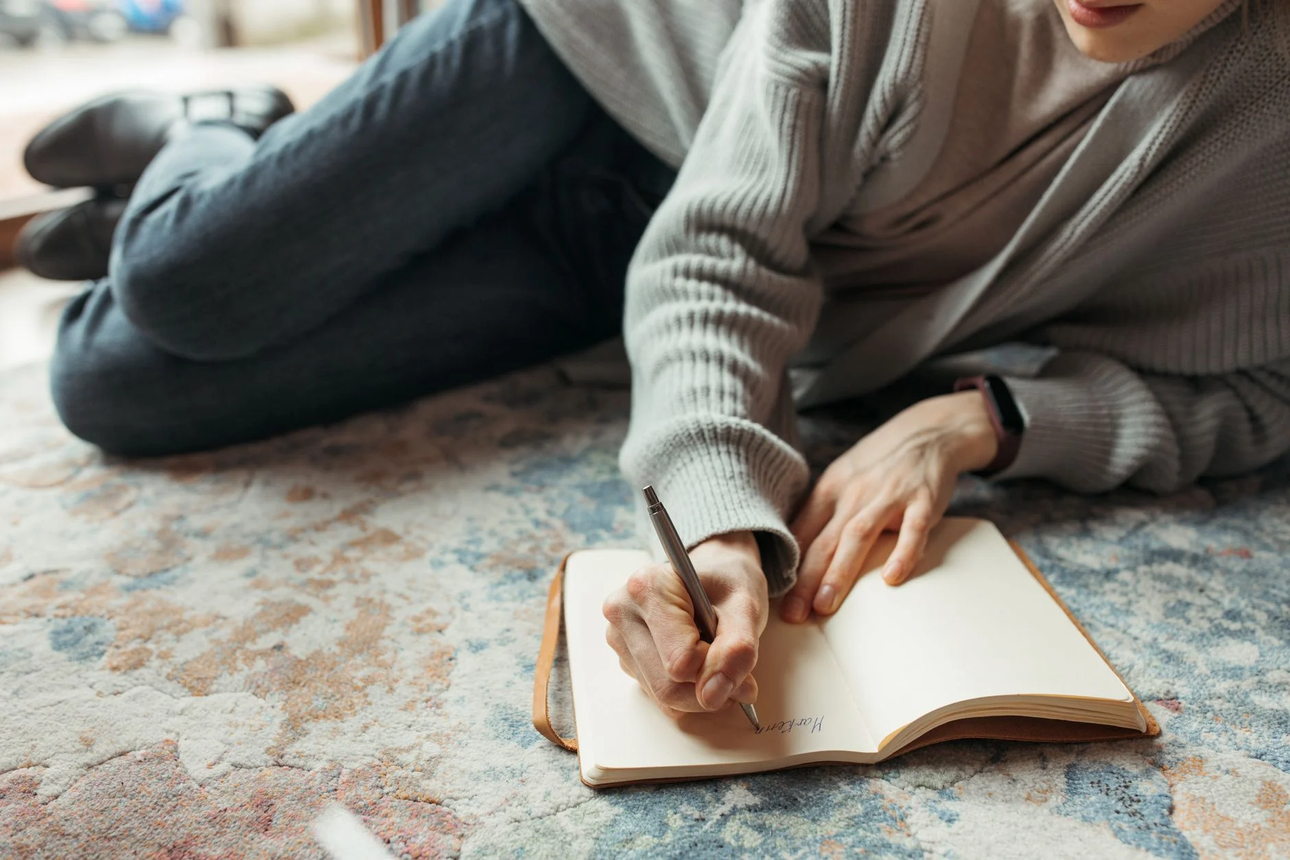A person lying on a rug writing in a notebook with a pen
