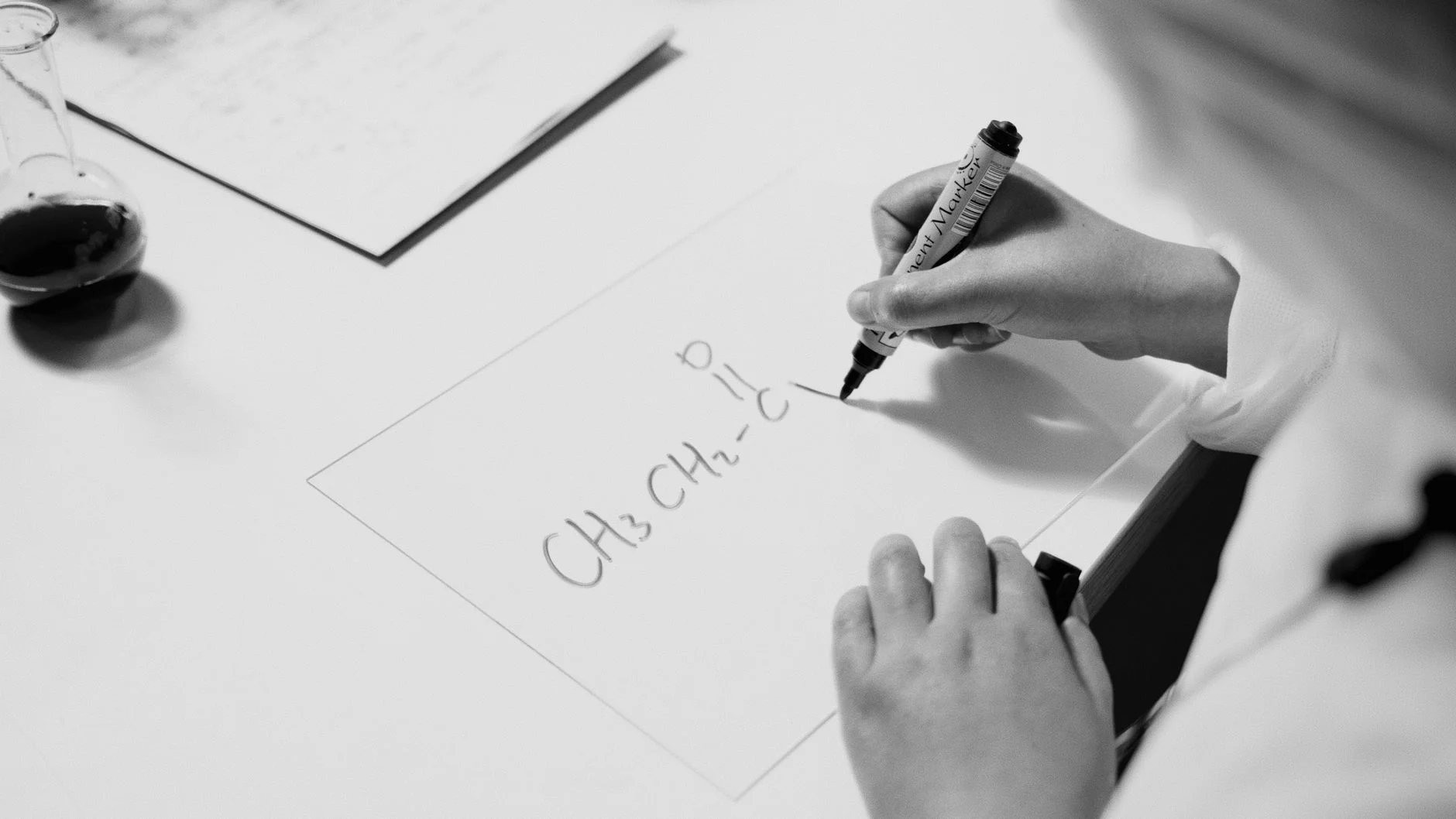 Black-and-white photo of a student’s hands writing a chemical formula with a marker on a card