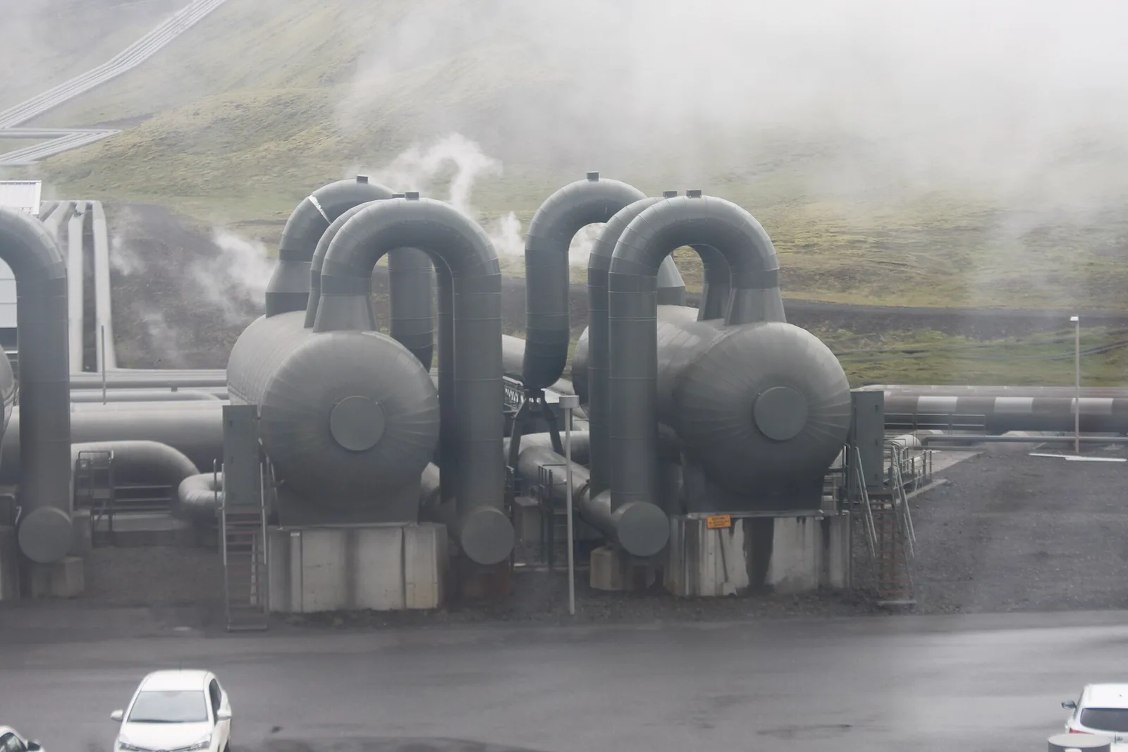 Large insulated pipework and steam vessels at Hellisheiði geothermal power station in Iceland, with steam drifting past