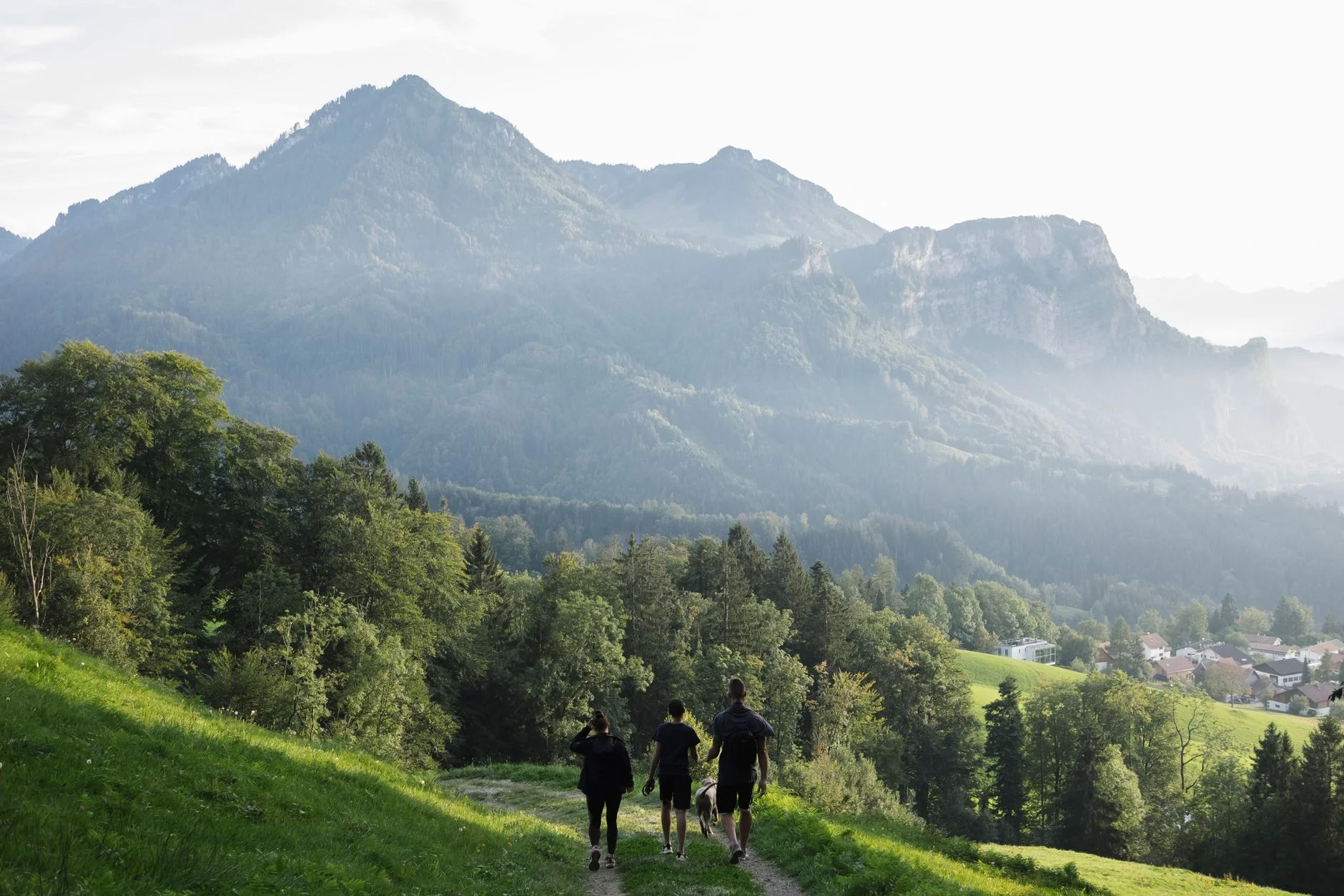 Three hikers walking down a grassy path towards a village with misty mountains behind