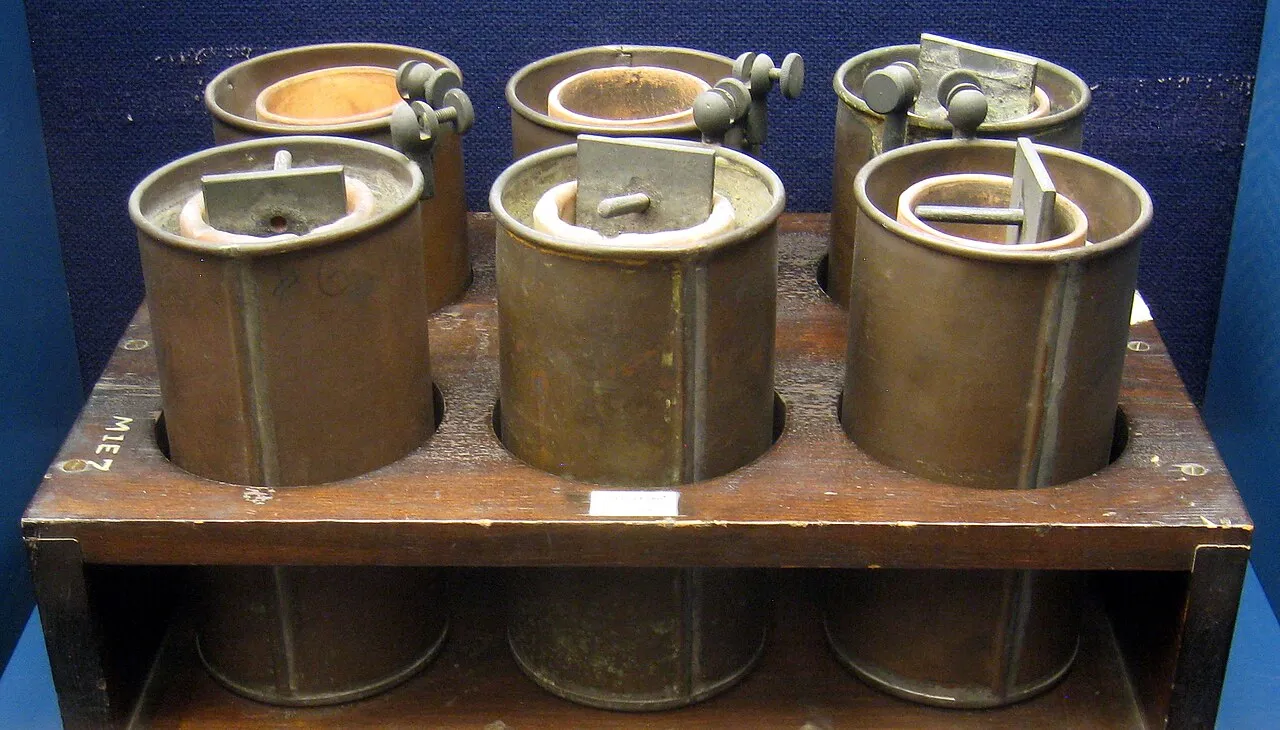 Six old copper Daniell cells in a wooden rack, each a copper cylinder with a terminal clamp, in a museum display