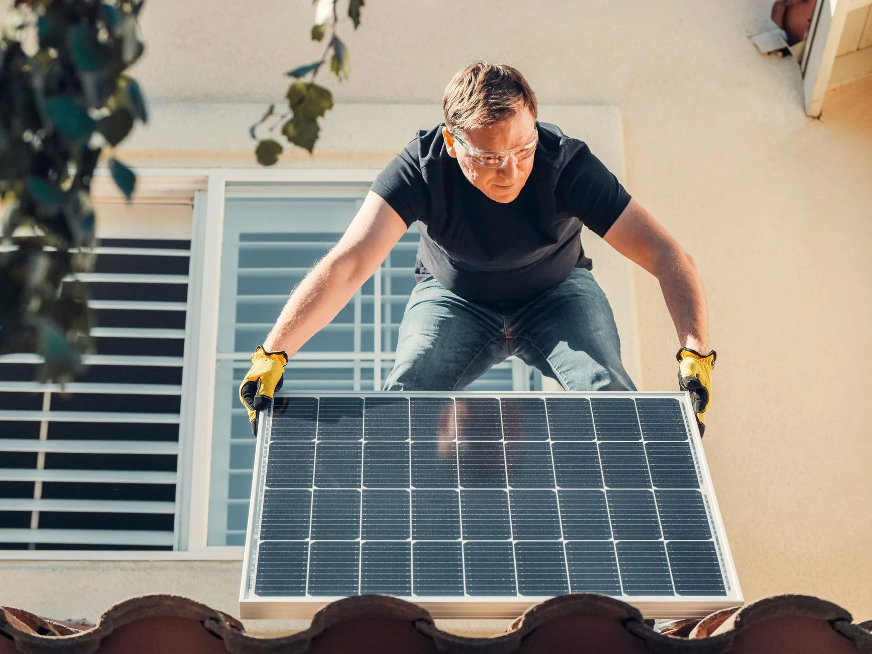 A man in safety glasses and gloves lifting a solar panel into place on a tiled house roof