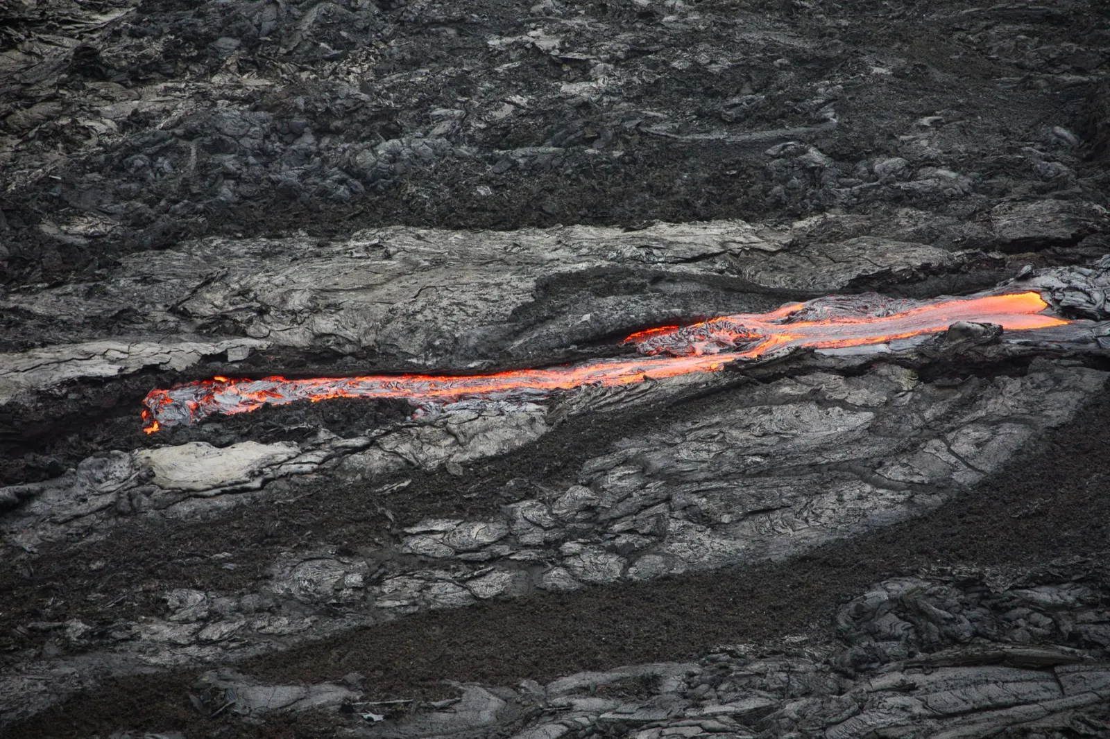 A thin stream of glowing orange lava flowing between dark cooled lava rock