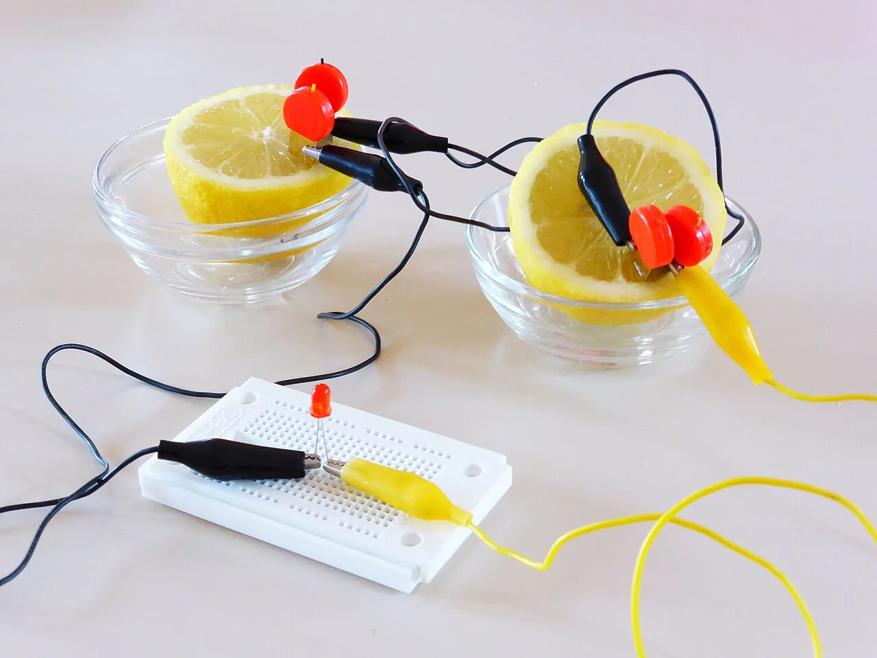 Two lemon halves in glass bowls wired in series with black and yellow crocodile leads to a red LED on a white breadboard