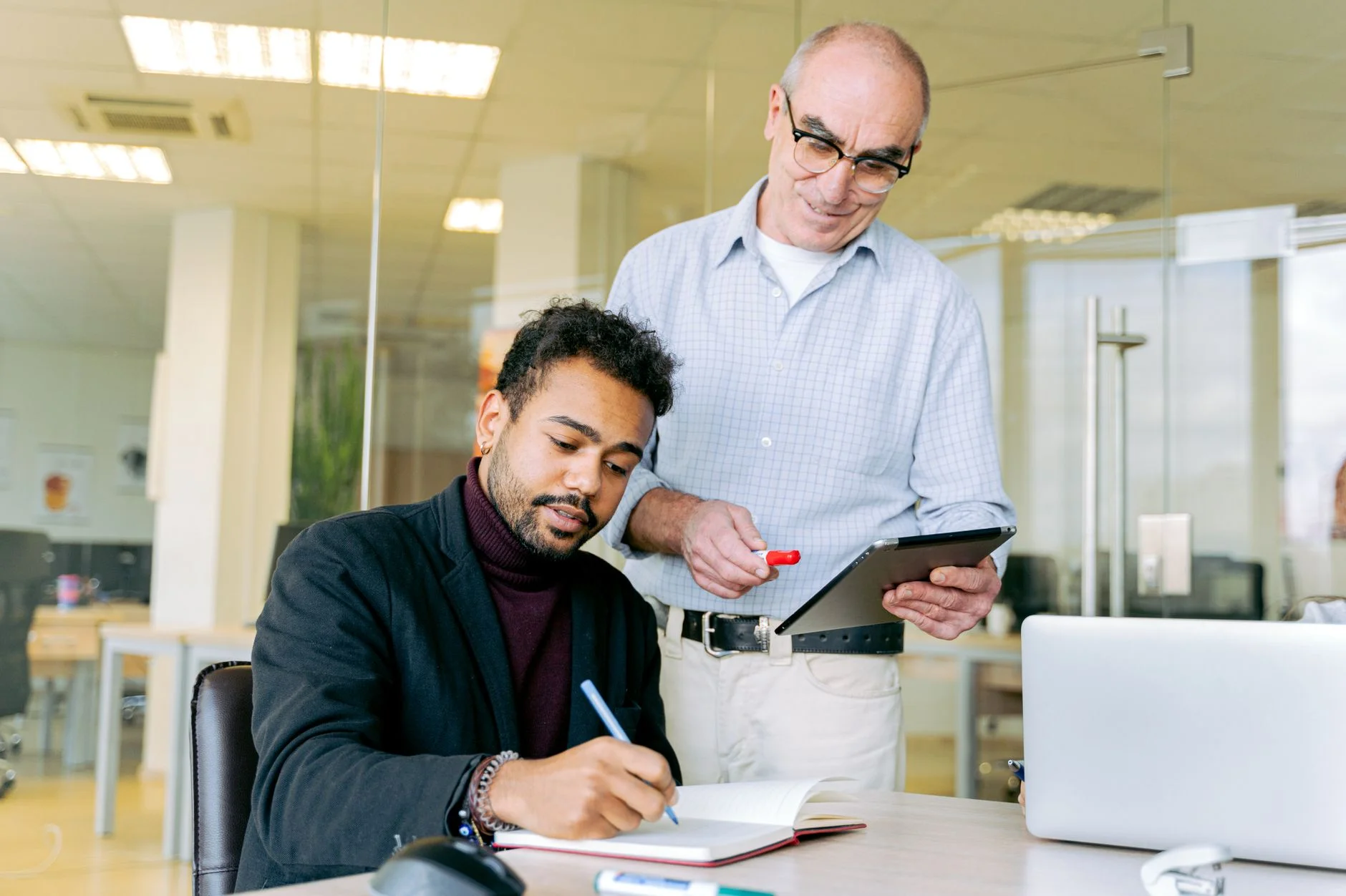 A manager standing beside a seated colleague, pointing at a tablet while the colleague takes notes