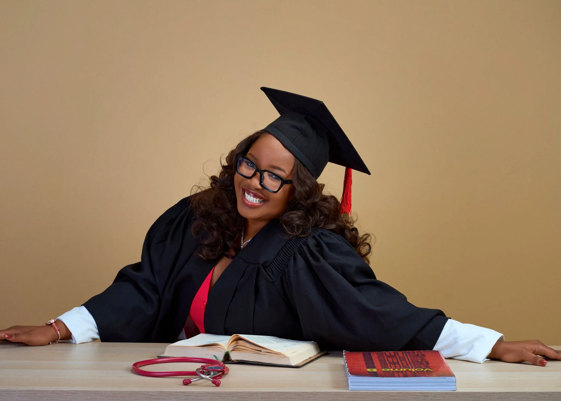 A smiling medical graduate in cap and gown with books and a stethoscope on the desk