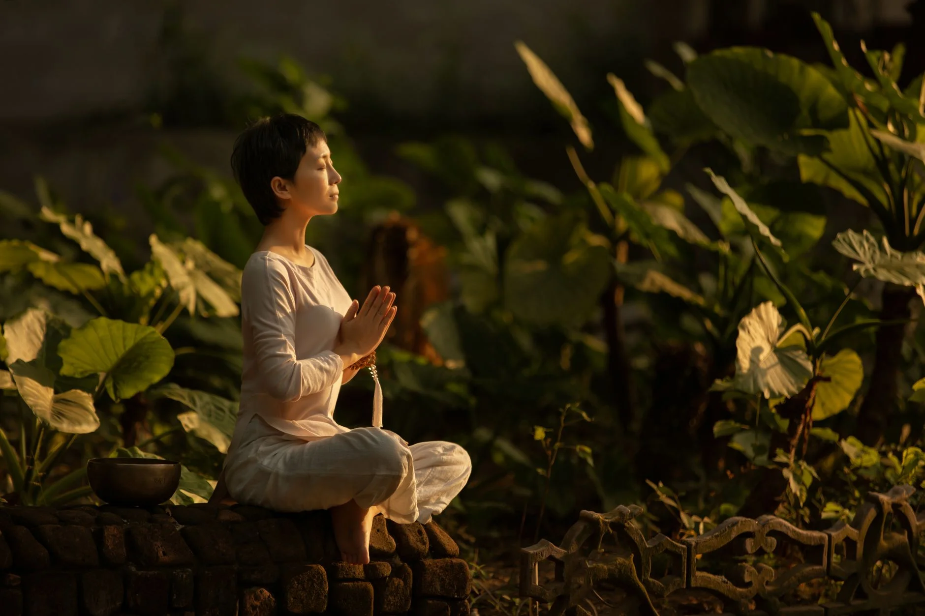 A woman sitting calmly with her hands together in a garden of large green leaves
