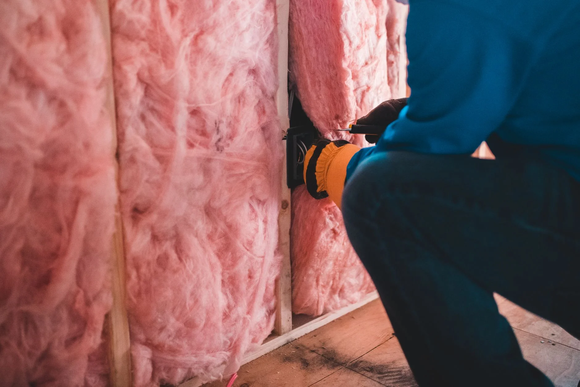 Worker kneeling beside wall bays packed with pink fibrous insulation between wooden studs