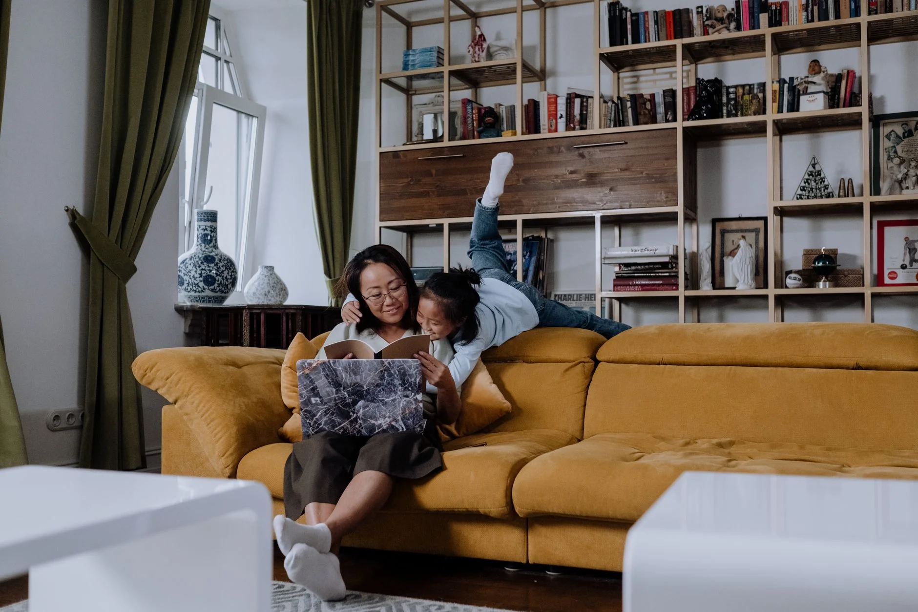 A mother and daughter reading a book together on a yellow sofa in a bright living room