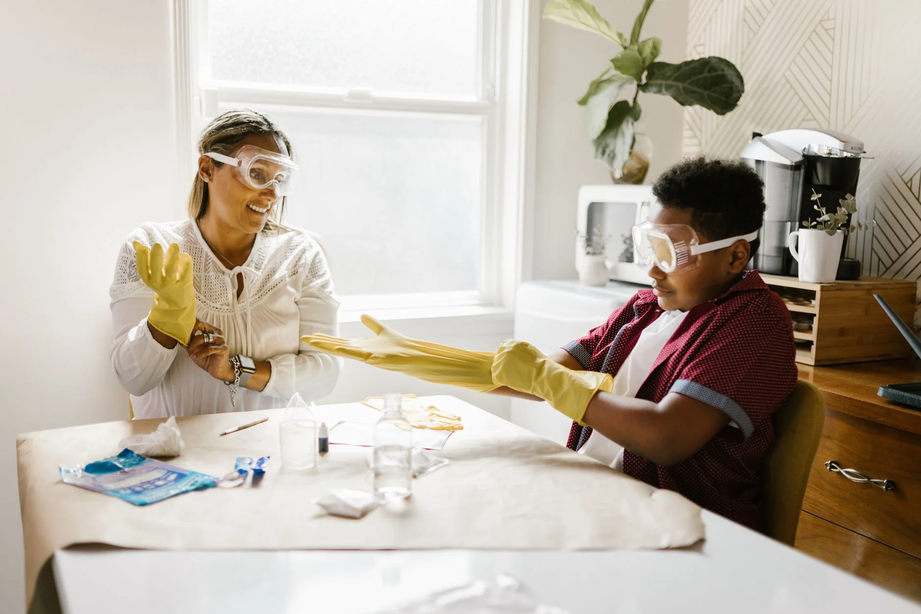 An adult and a child wearing safety goggles and rubber gloves doing a science experiment together at a kitchen table