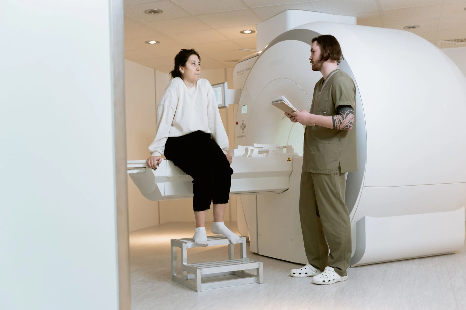 A radiographer talking with a patient sitting on the table of an MRI scanner in a bright scanning room
