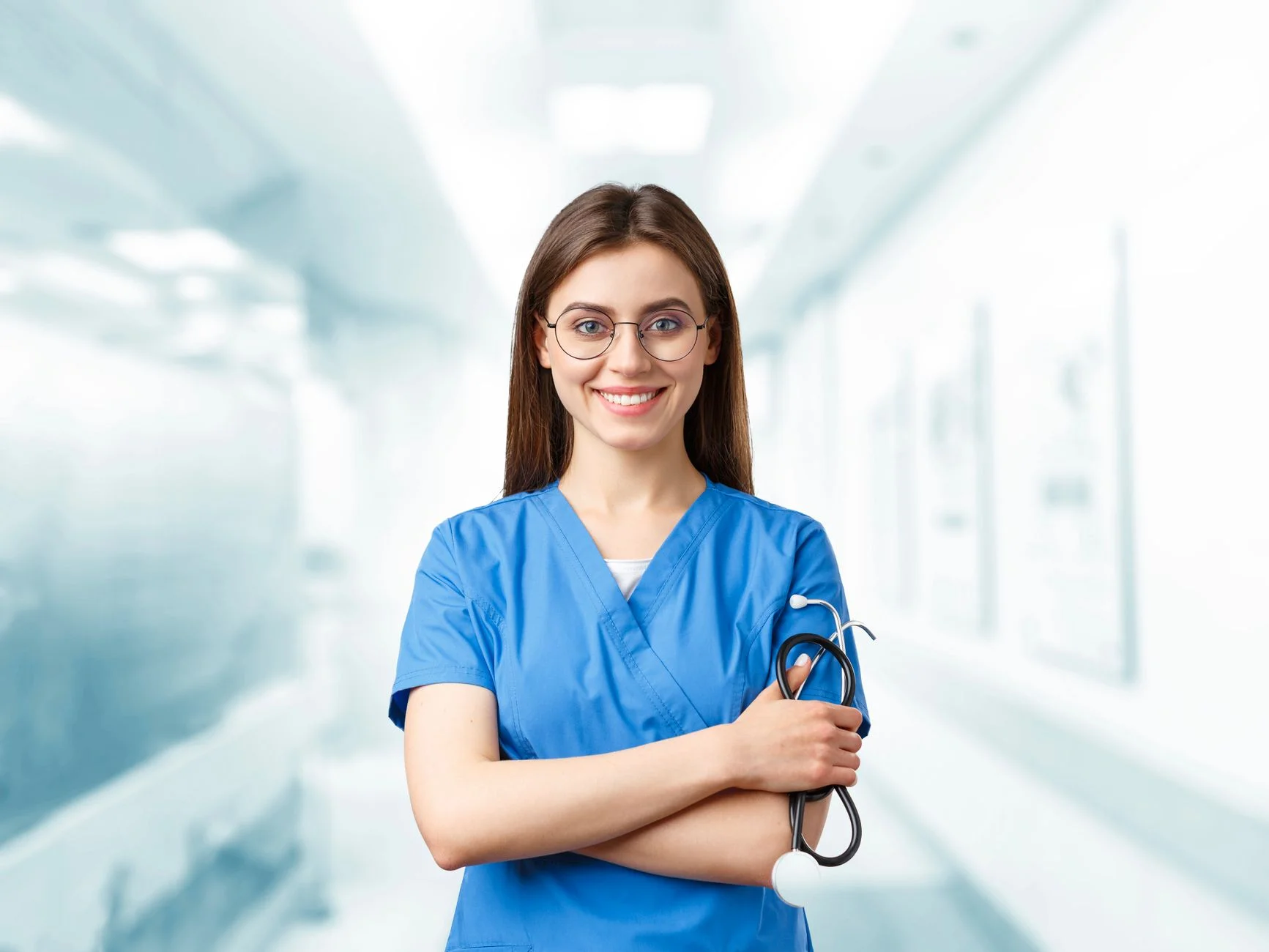 A smiling nurse in blue scrubs holding a stethoscope in a bright hospital corridor