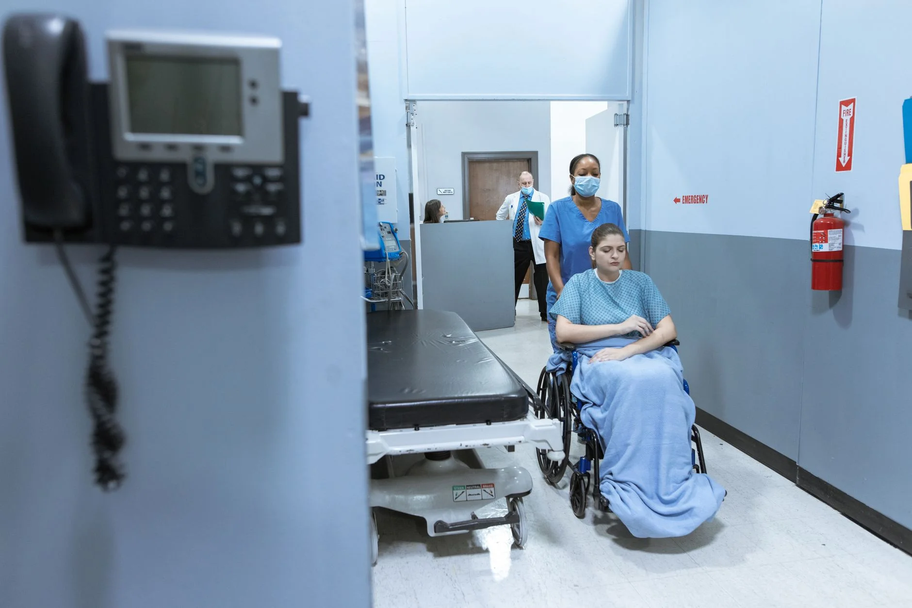 A nurse in scrubs pushing a patient in a wheelchair along a hospital corridor