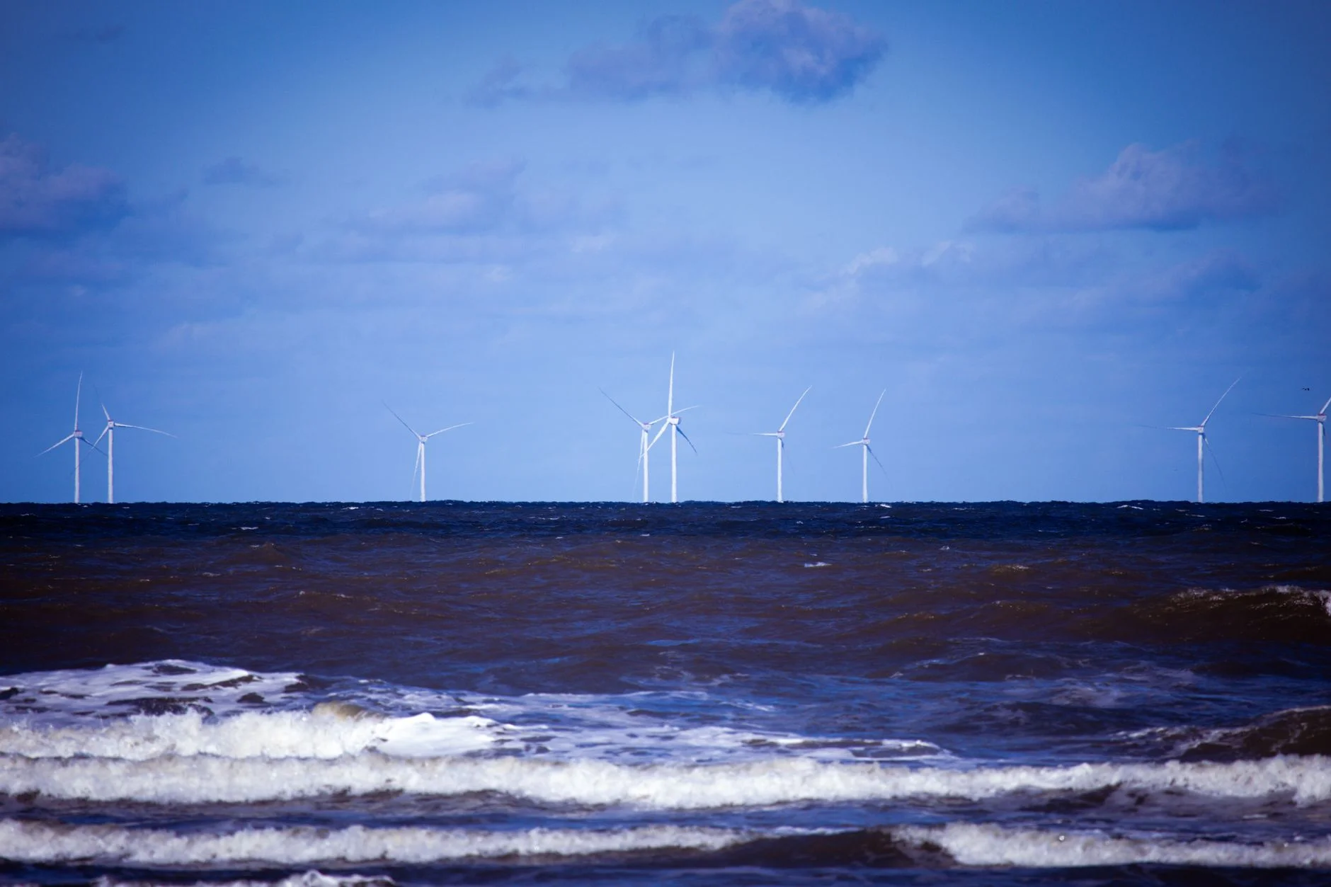 A line of offshore wind turbines on the horizon beyond rough blue sea and breaking waves