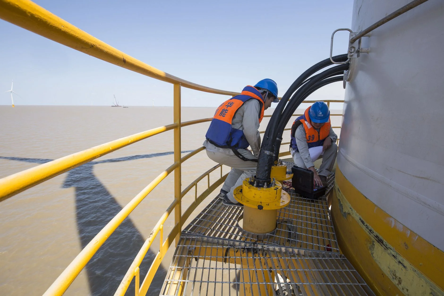 Two technicians in hard hats and life jackets working on cables at the base of an offshore wind turbine tower