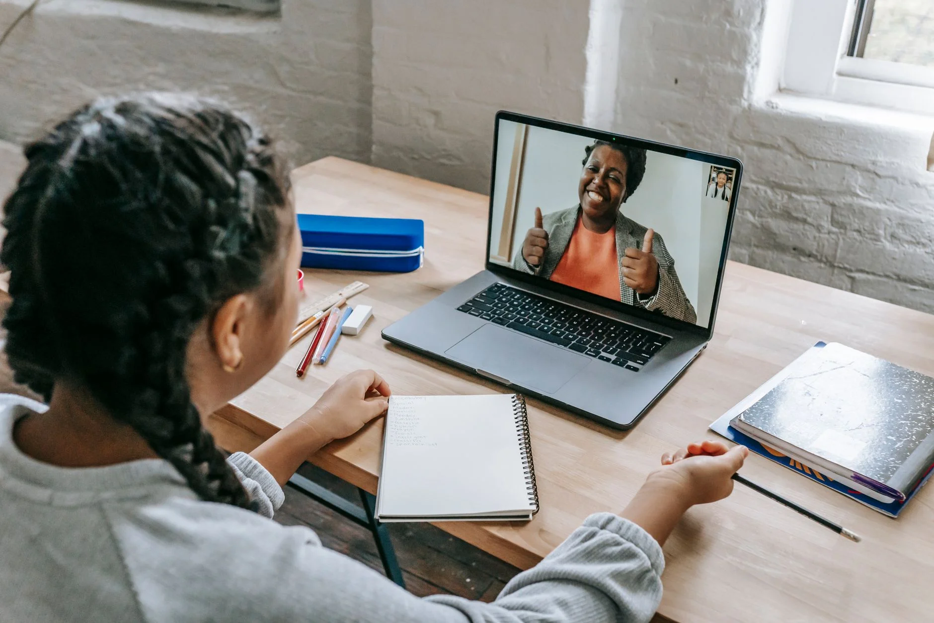 A student with braids sitting at a desk with a notebook while a smiling tutor gives a thumbs up on a laptop video call