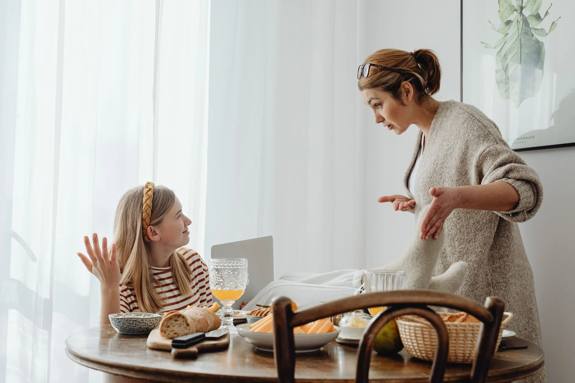 A mother standing and gesturing at a teenage daughter seated at a breakfast table in a tense exchange
