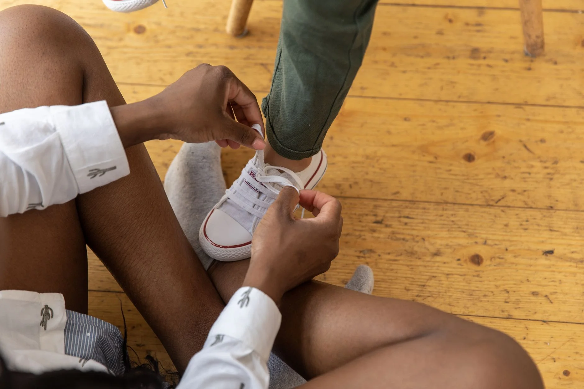 An adult’s hands tying a child’s white trainer laces on a wooden floor