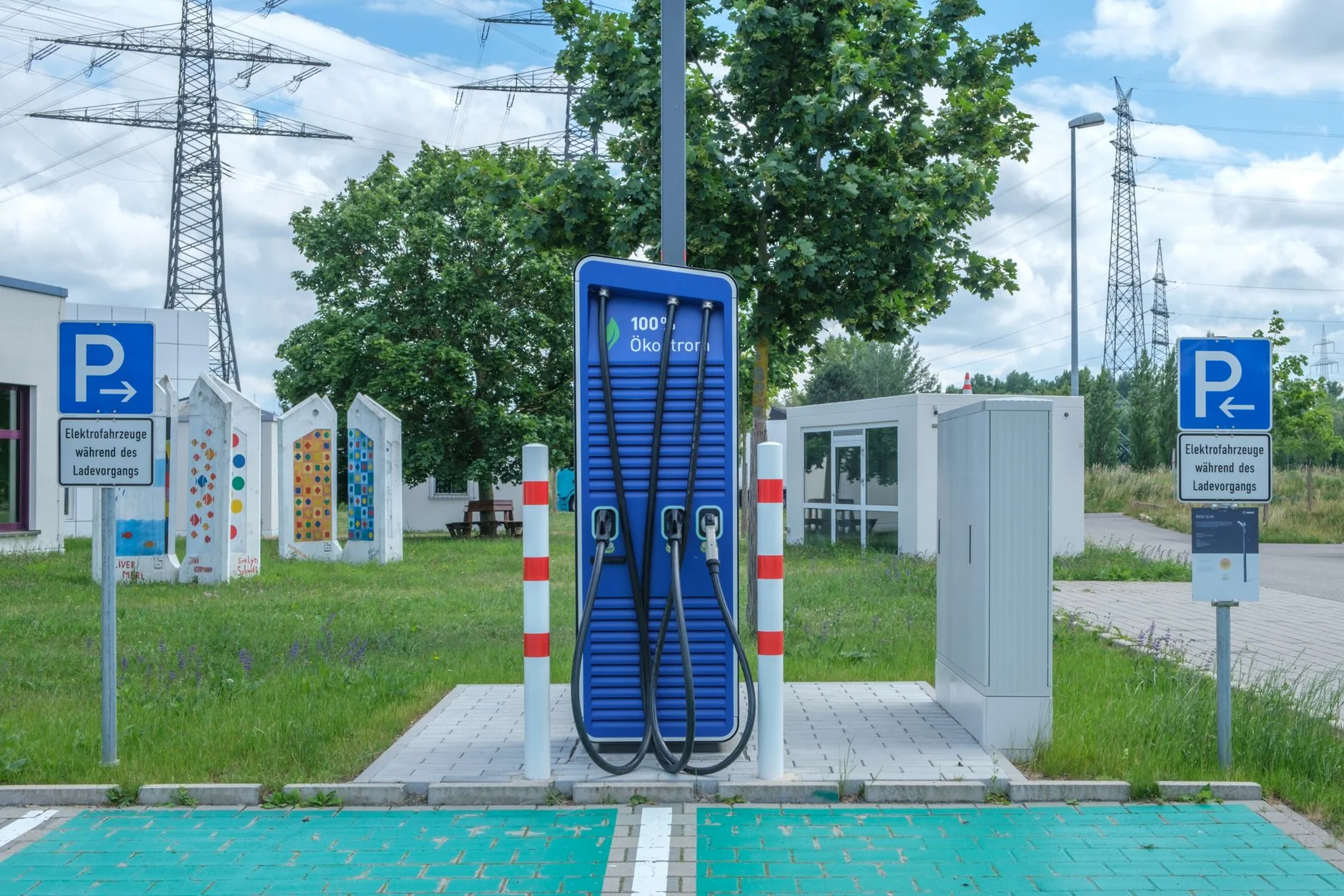 A blue public rapid charger with three tethered cables on a paved bay, with green-painted parking spaces and electricity pylons behind